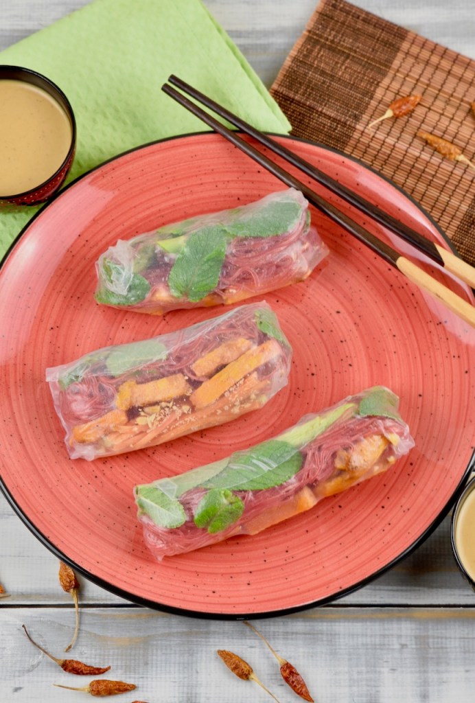 Drei Sommerrollen mit grünem Spargel und Süßkartoffel auf einem roten Teller. Auf dem Teller liegen auch Stäbchen. Im Hintergrund sieht man zwei Schälchen mit Erdnusssauce.