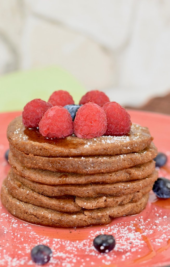 Glutenfreie Pfannkuchen gestapelt auf roten Teller, garniert mit Himbeeren, Blaubeeren, Ahornsirup und Puderzucker. Hintergrund hell.