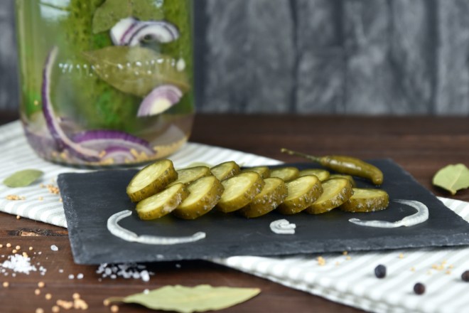 Fermentierte Gurken in einem Einmachglas. Davor liegen die Senfgurken angeschnitten auf einer Schieferplatte.