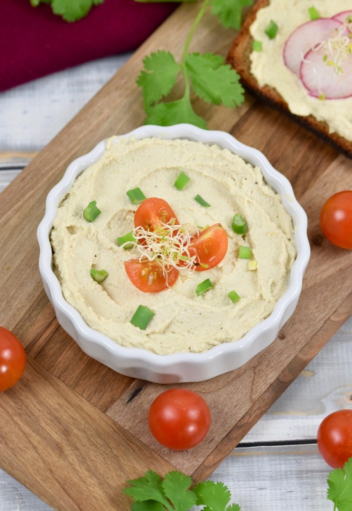 Veganer Frischkäse in weißer Schale, die auf einem Holzbrettchen steht. Der Frischkäse ist mit Vierteln von Cocktailtomaten, Frühlingszwiebel und Sprossen garniert. Im Hintergrund liegt eine Scheibe Brot mit Frischkäse und Radieschen.
