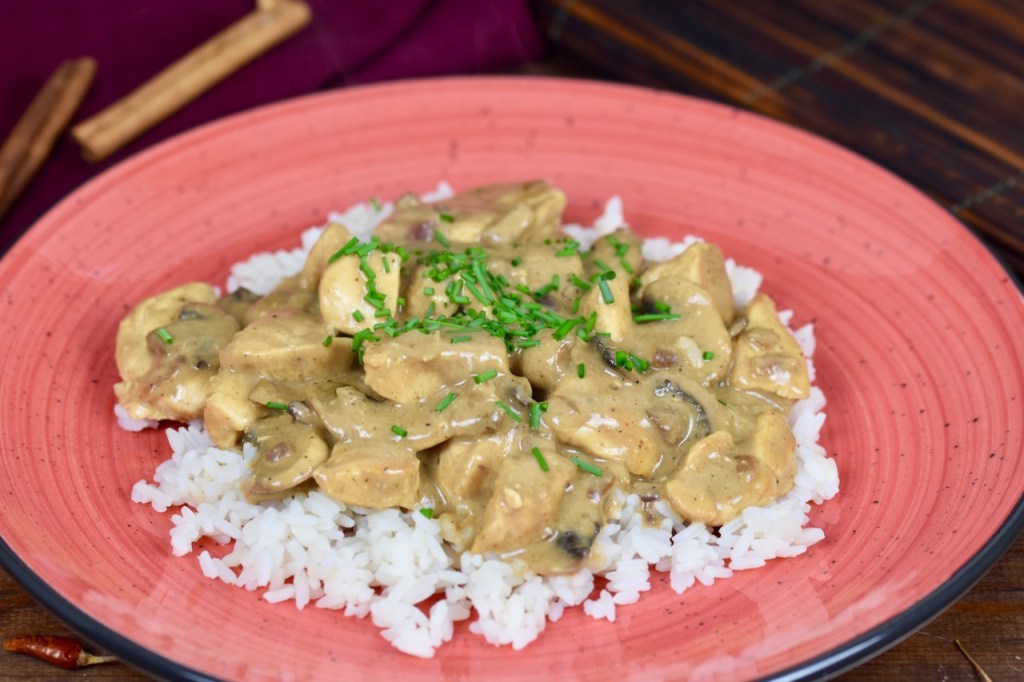 Hähnchen-Geschnetzeltes mit Pilzen auf Reis auf einem roten Teller, dekoriert mit Schnittlauchröllchen. Hintergrund dunkel