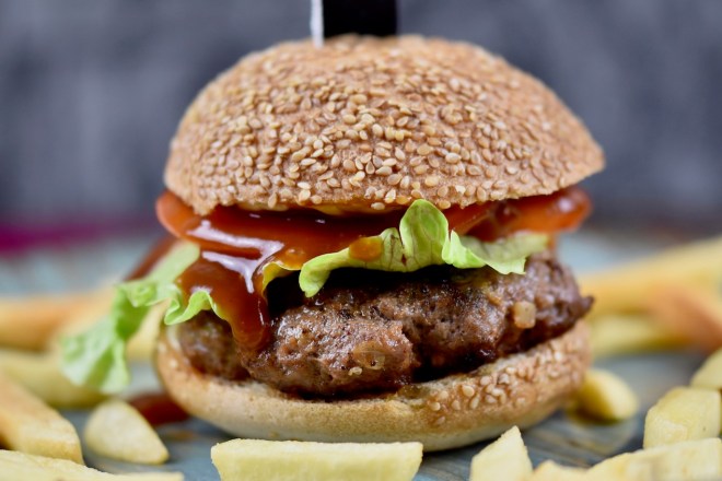 Pljescavica Burger aufgespießt mit einem Messer auf einem bläulichen Teller. Drumherum liegen Pommes frites. Dunkler Hintergrund.
