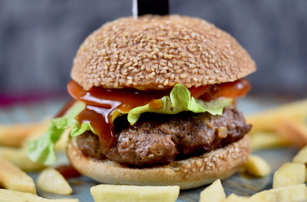 Pljescavica Burger aufgespießt mit einem Messer auf einem bläulichen Teller. Drumherum liegen Pommes frites. Dunkler Hintergrund.
