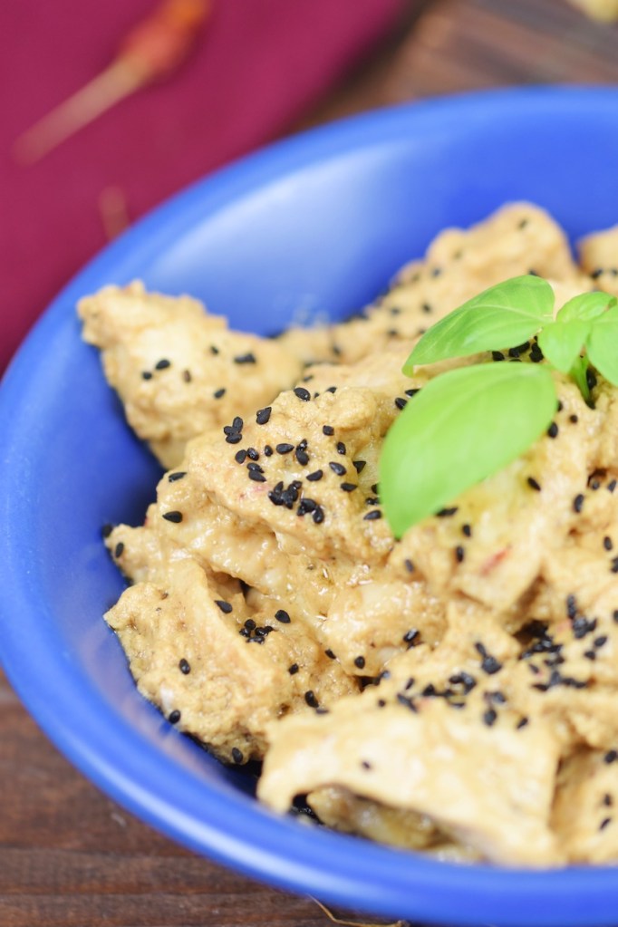 Hähnchen-Mezze in Tahini-Kokossauce in einer blauen Schale garniert mit Basilikum. Hintergrund dunkel.