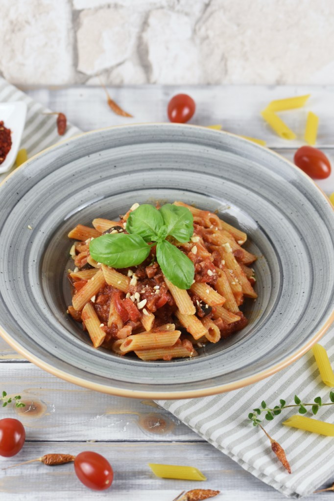 Penne Arrabiata mit Paprika und Oliven auf einem grauen Teller. Hintergrund hell.