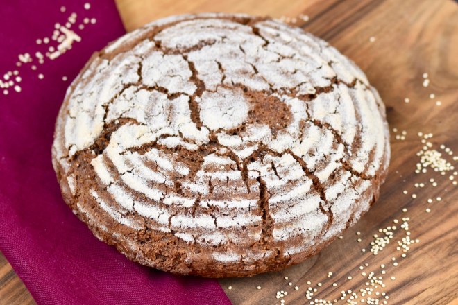 glutenfreies Krustenbrot in rund auf Holzbrett. Dekoriert ist es mit einer weinroten Stoffserviette und groben Salzkörnern.