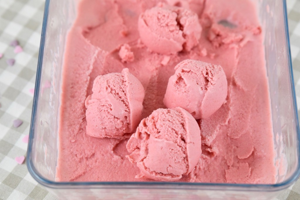 Erdbeereis in einer eckigen Auflaufform aus Glas. Auf dem gefrorenen Eis liegen vier Eiskugeln.