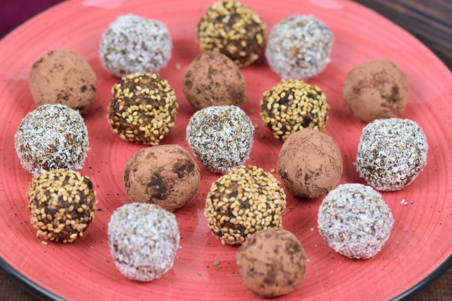 Glücksbällchen - Bliss Balls - ummantelt mit Kakaopuler, Kokosraspeln oder Sesam auf rotem Teller.