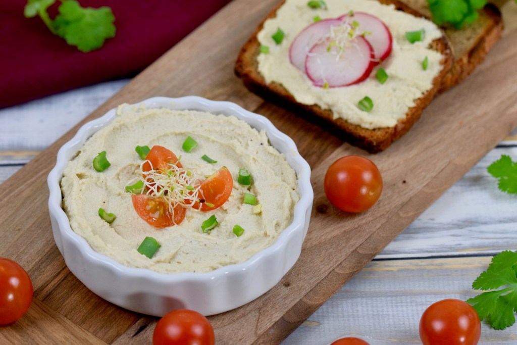 Veganer Frischkäse in weißer Schale, die auf einem Holzbrettchen steht. Der Frischkäse ist mit Vierteln von Cocktailtomaten, Frühlingszwiebel und Sprossen garniert. Im Hintergrund liegt eine Scheibe Brot mit Frischkäse und drei Scheiben Radieschen.