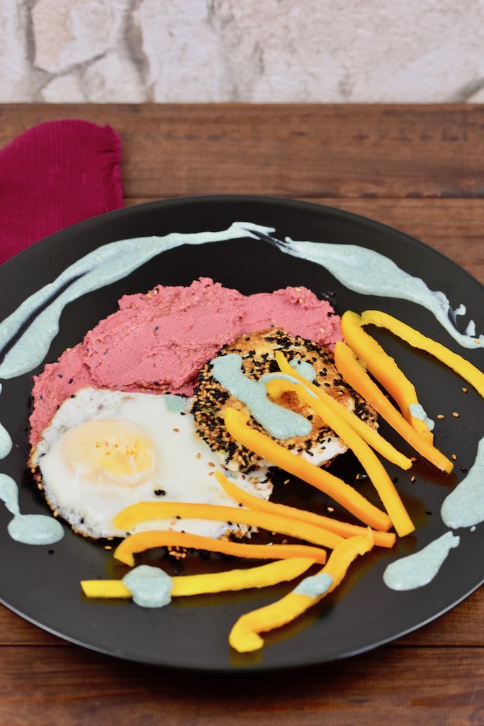 Knusprige Spiegeleier mit einer Kruste aus Sesam und Schwarzkümmelsamen mit pinkem Rote Beete Hummus und grüner Tahini-Sauce. Dazu gibt es gelbe Paprikastreifen Angerichtet ist alles auf einem schwarzen Teller.