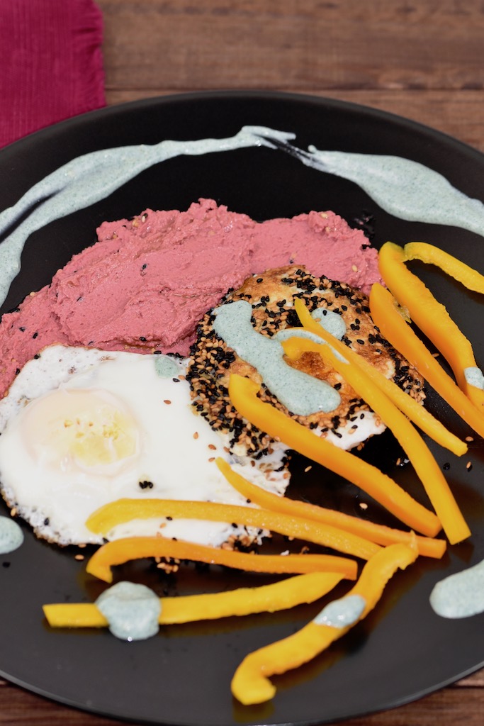 Knusprige Spiegeleier mit einer Kruste aus Sesam und Schwarzkümmelsamen mit pinkem Rote Beete Hummus und grüner Tahini-Sauce. Dazu gibt es gelbe Paprikastreifen Angerichtet ist alles auf einem schwarzen Teller.