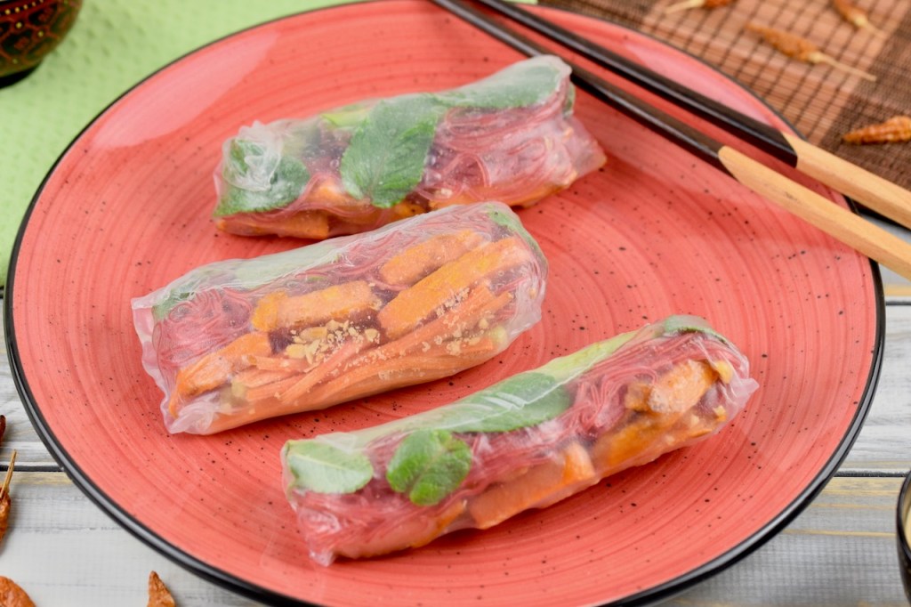 Drei Sommerrollen mit grünem Spargel und Süßkartoffel auf einem roten Teller. Auf dem Teller liegen auch Stäbchen.