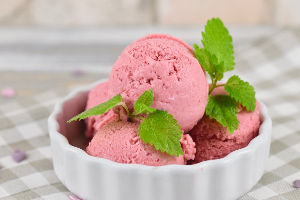 Veganes Erdbeereis selber machen - 4 Kugeln Erdbeereis in einer weißen Schale garniert mit Zitronenmelisse. Hintergrund hell.