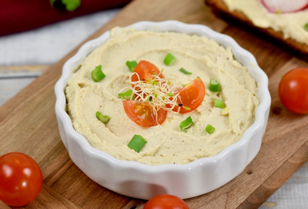 Veganer Frischkäse in weißer Schale, die auf einem Holzbrettchen steht. Der Frischkäse ist mit Vierteln von Cocktailtomaten, Frühlingszwiebel und Sprossen garniert. Nahaufnahme