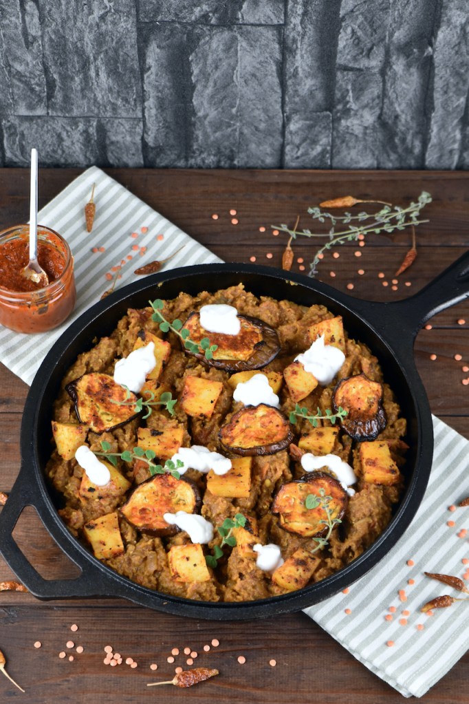 Linsen mit Harissa Kartoffeln und Auberginen mit ein paar Klecksen Joghurt dekoriert in einer schwarzen Pfanne aus Gusseisen.