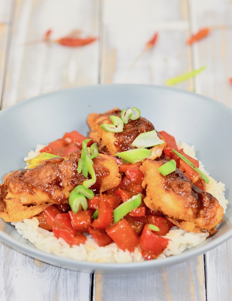 Gebackenes Hoisin-Hähnchen auf Paprikagemüse mit Reis in hellgrauem Teller. Der Teller steht auf einer weißen Holzfläche.