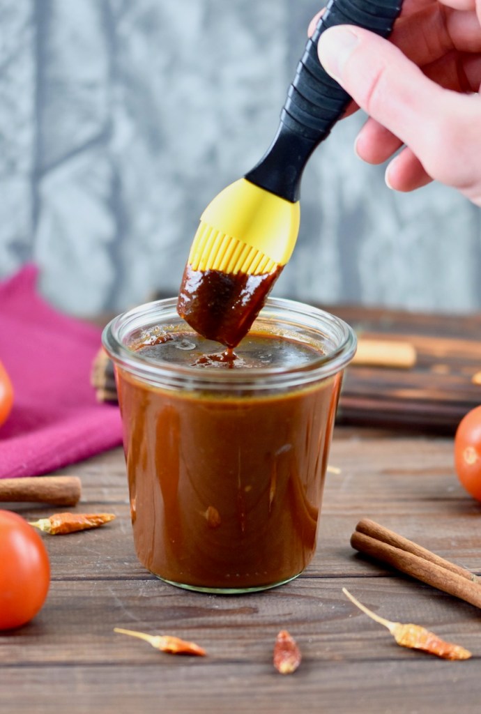 Rauchige BBQ-Sauce in einem Glas. Ein Pinsel mit gelben Silikonborsten wird in die Sauce getunkt. Hintergrund dunkel.