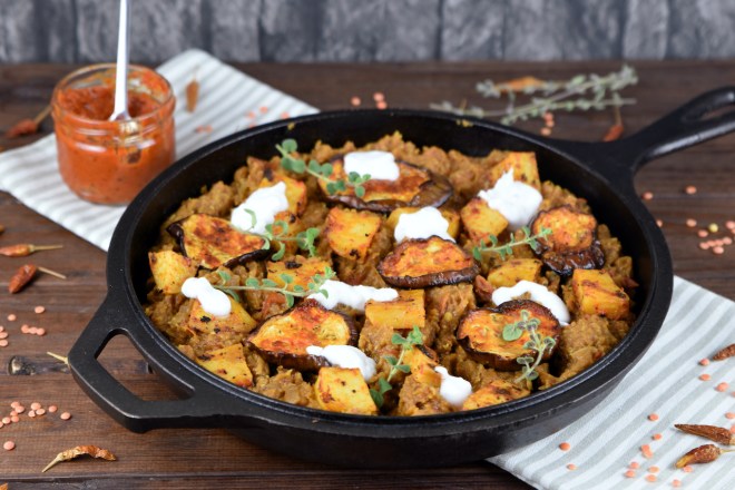 Linsen mit Harissa Kartoffeln und Auberginen mit ein paar Klecksen Joghurt dekoriert in einer schwarzen Pfanne aus Gusseisen.