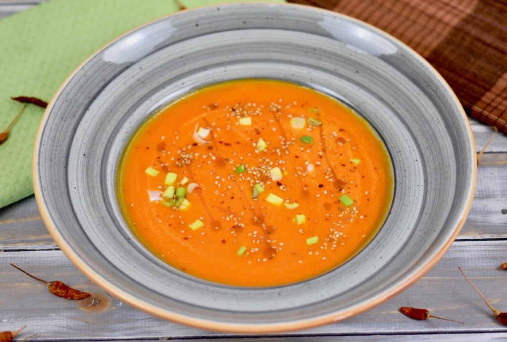 Karottensuppe mit Ingwer und Orange in grauem Teller. Garniert mit Sesamsamen, Sesamöl und Frühlingszwiebeln. Hintergrund hell.