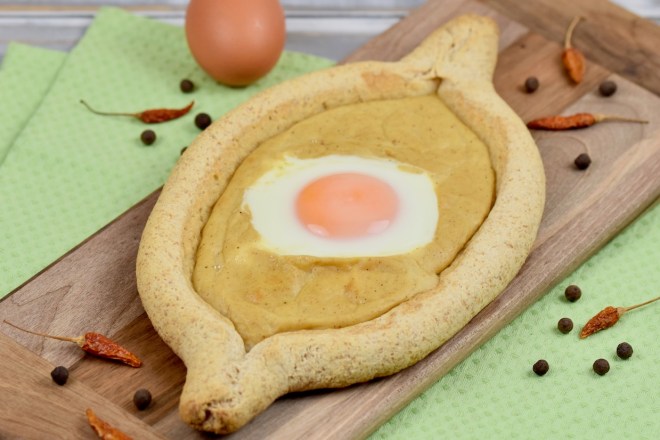 Chatschapuri mit Ei und Senfsauce auf länglichem Holzbrettchen. Heller Hintergrund dekoriert mit einem Ei, getrockneten Chilischoten und Pimentkörnern.