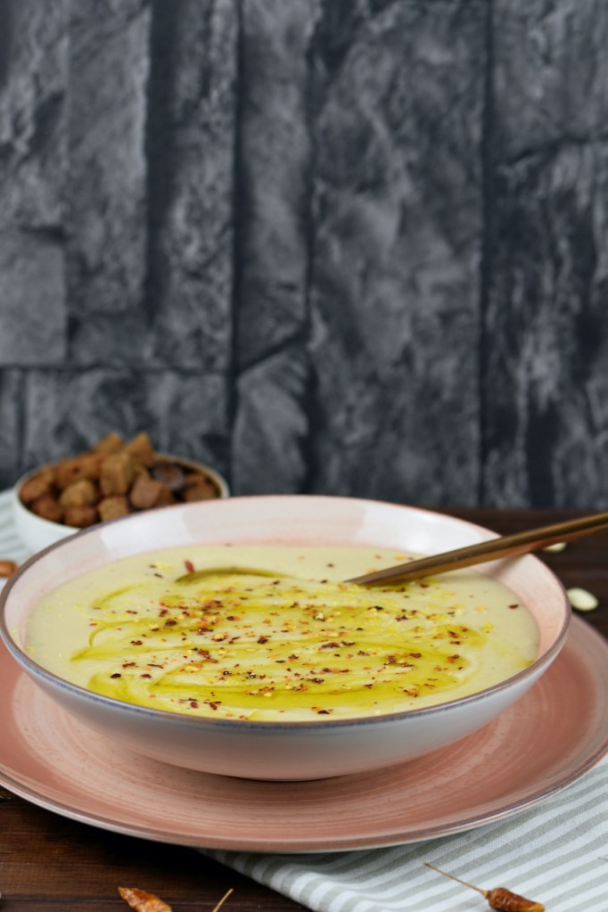 Cremige Blumenkohlsuppe in einem rosa Teller. Im Hintergrund stehen die Croûtons.