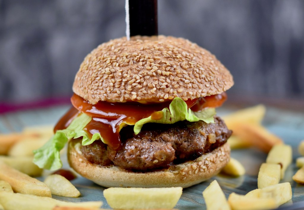 Pljescavica Burger aufgespießt mit einem Messer auf einem bläulichen Teller. Drumherum liegen Pommes frites. Dunkler Hintergrund.