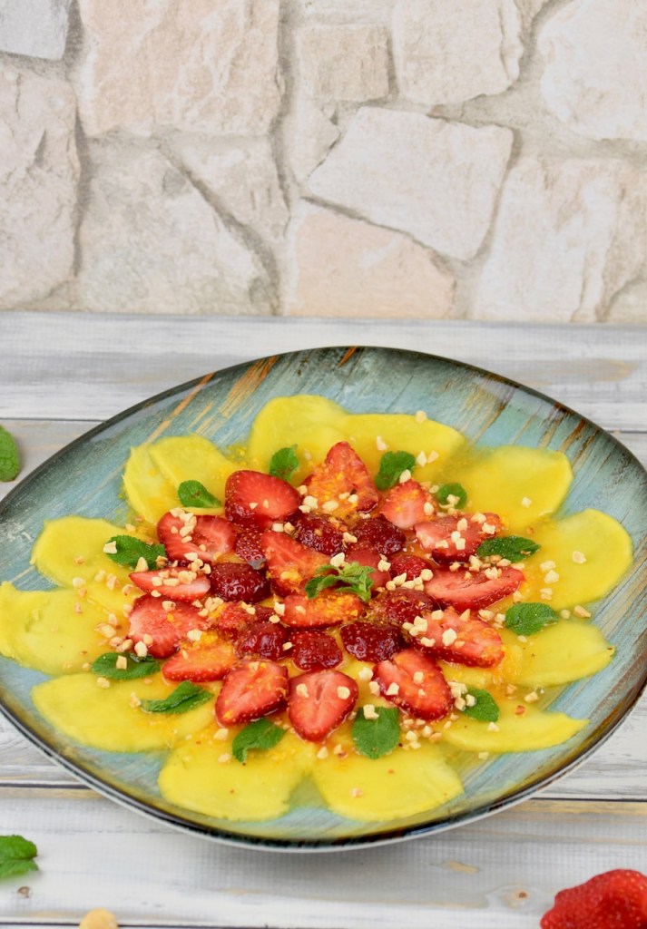 Erdbeer-Mango-Carpaccio mit Minze und Orangen-Dressing auf türkisem Teller garniert mit Minze und Haselnusskernen. Hintergrund weiß.