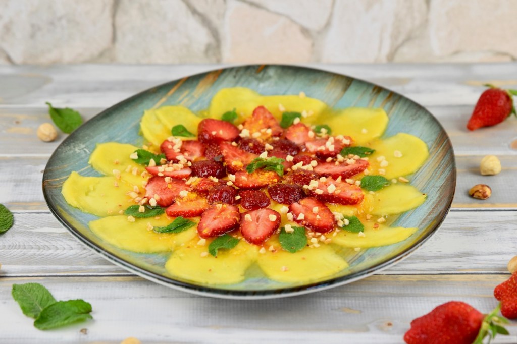 Erdbeer-Mango-Carpaccio mit Minze und Orangen-Dressing auf türkisem Teller garniert mit Minze und Haselnusskernen. Hintergrund weiß.