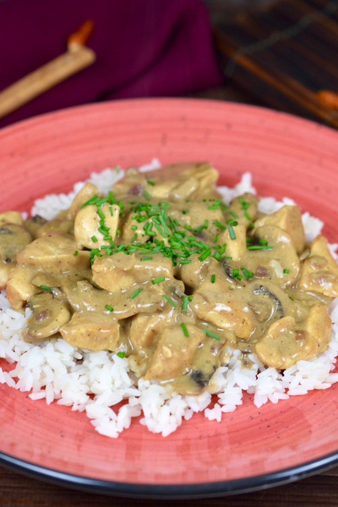 Hähnchen-Geschnetzeltes mit Pilzen auf Reis auf einem roten Teller, dekoriert mit Schnittlauchröllchen. Hintergrund dunkel