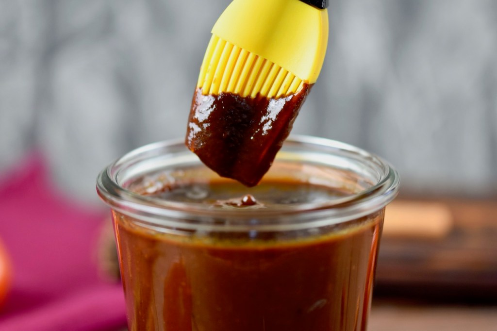 Rauchige BBQ-Sauce in einem Glas. Ein Pinsel mit gelben Silikonborsten wird in die Sauce getunkt. Hintergrund dunkel.