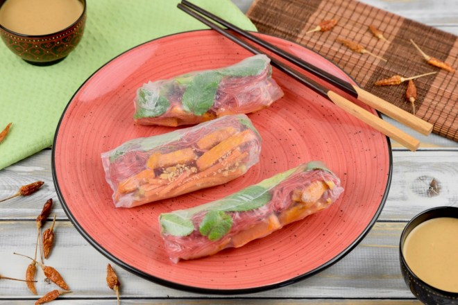 Drei Sommerrollen mit grünem Spargel und Süßkartoffel auf einem roten Teller. Auf dem Teller liegen auch Stäbchen. Im Hintergrund sieht man zwei Schälchen mit Erdnusssauce.