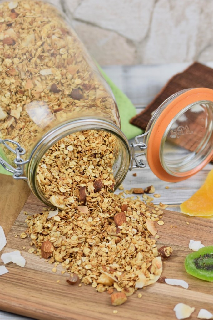Knuspriges Granola, das aus einem liegenden Glas geschüttet wurde. Das ausgeschüttete Granola liegt auf einem Holzbrett und der Hintergrund ist hell.