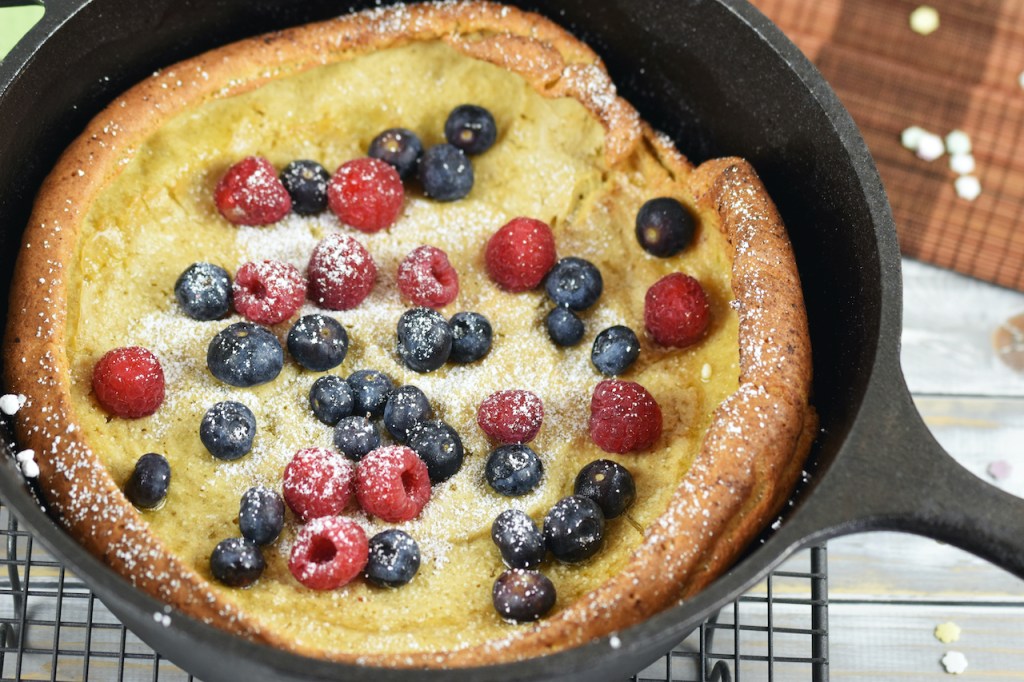 Glutenfreier Dutch Baby im schwarzen Gusseisentopf, dekoriert mit Himbeeren, Blaubeeren und Puderzucker. Hintergrund hell.