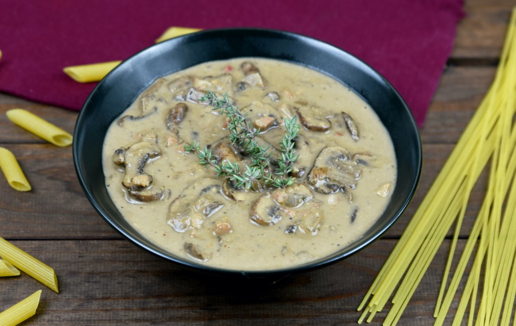 Cremige Pilzsauce aus Cashewkernen in einer schwarzen Schüssel. Drumherum liegen italienische Pasta. Hintergrund dunkel.