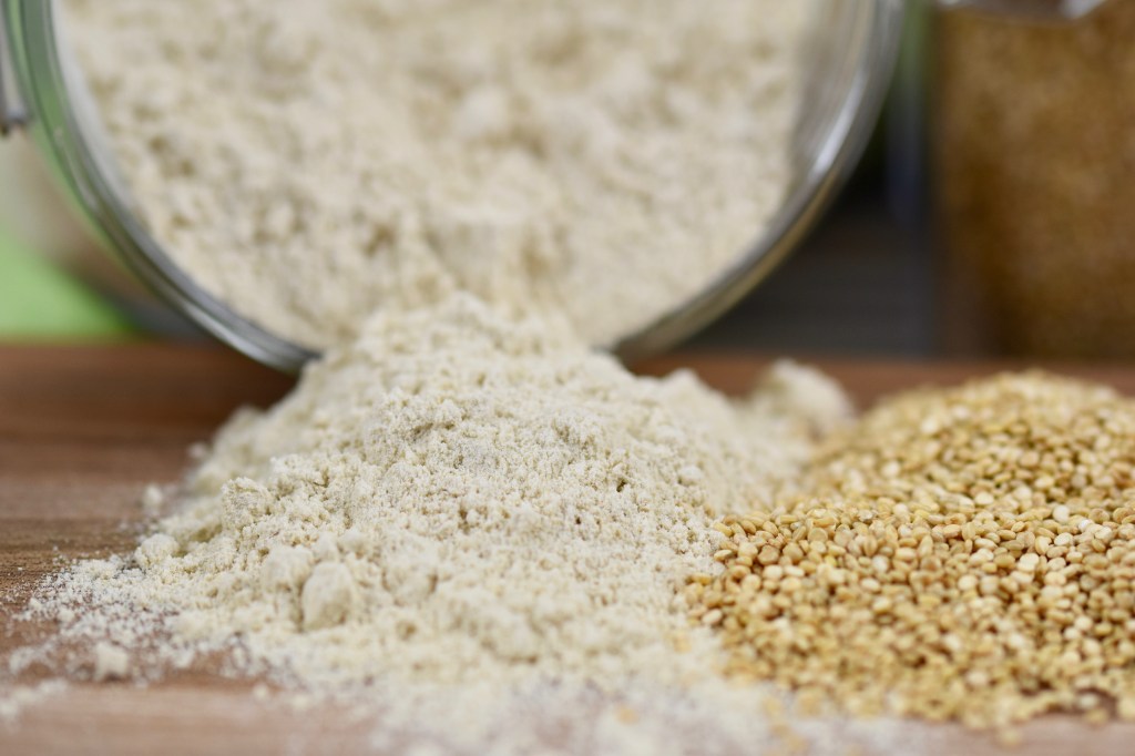 Quinoamehl aus einem Glas auf ein Holzbrett geschüttet. Daneben liegen Quinoakörner.