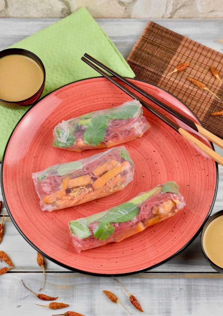 Drei Sommerrollen mit grünem Spargel und Süßkartoffel auf einem roten Teller. Auf dem Teller liegen auch Stäbchen. Im Hintergrund sieht man zwei Schälchen mit Erdnusssauce.