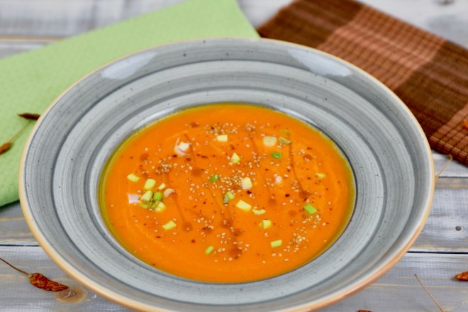 Karottensuppe mit Ingwer und Orange in grauem Teller. Garniert mit Sesamsamen, Sesamöl und Frühlingszwiebeln. Hintergrund hell.
