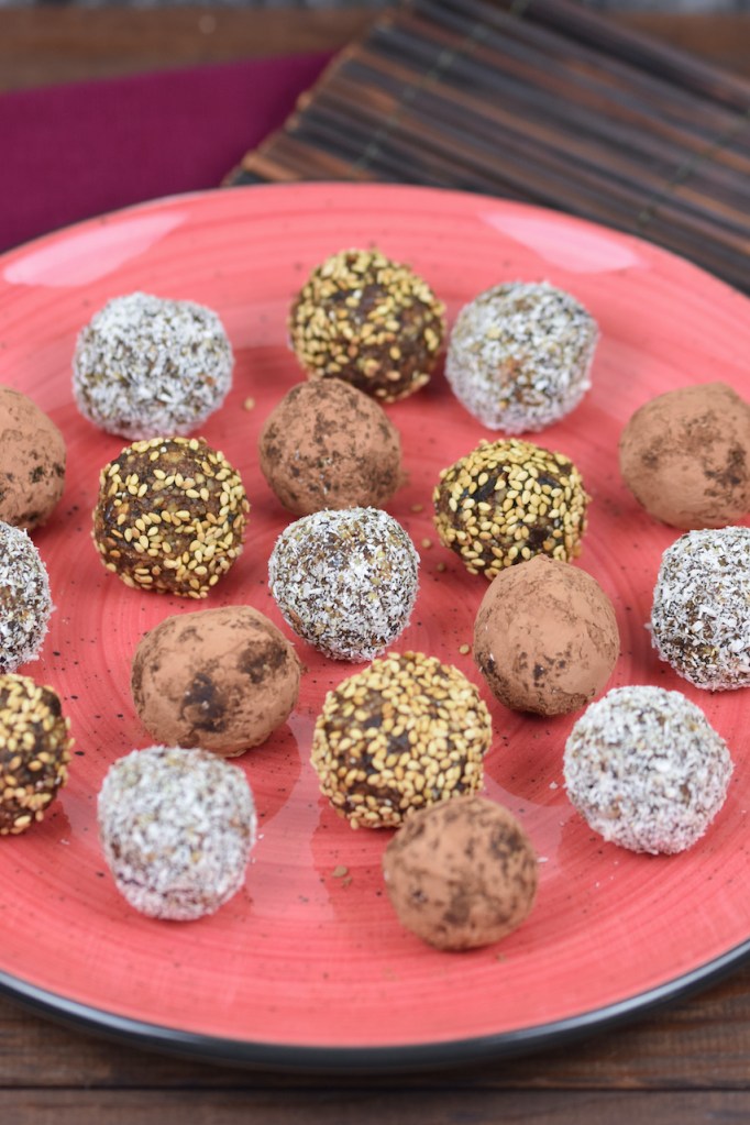 Glücksbällchen - Bliss Balls - ummantelt mit Kakaopuler, Kokosraspeln oder Sesam auf rotem Teller.