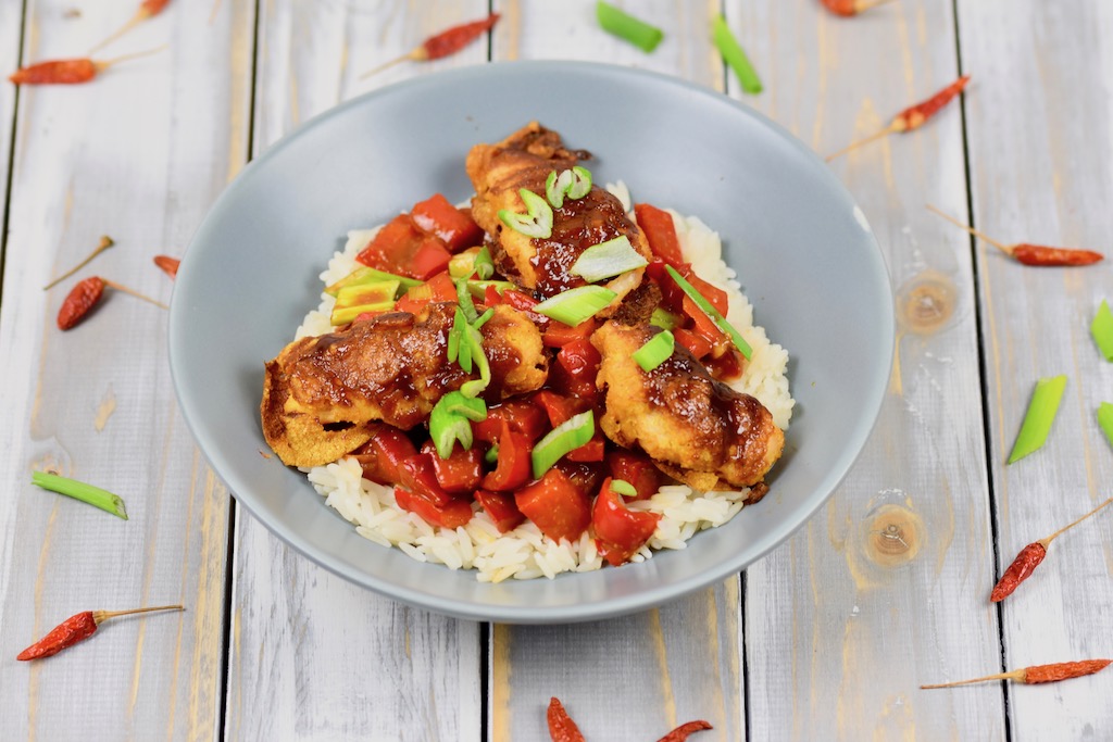 Gebackenes Hoisin-Hähnchen auf Paprikagemüse mit Reis in hellgrauem Teller. Der Teller steht auf einer weißen Holzfläche.