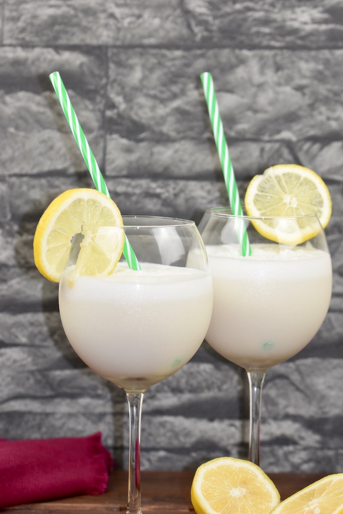 Tropische Kokoslimonade in zwei Weingläsern garniert mit einer Zitronenscheibe und einem Strohhalm. Hintergrund dunkel.