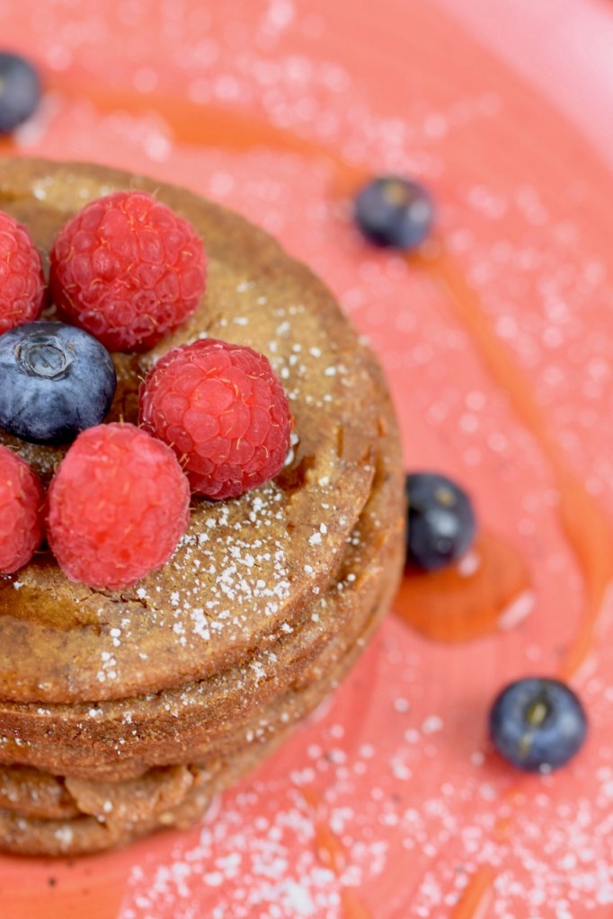Glutenfreie Pfannkuchen gestapelt auf roten Teller, garniert mit Himbeeren, Blaubeeren, Ahornsirup und Puderzucker. Es ist nur der halbe Pfannkuchenturm zu sehen.