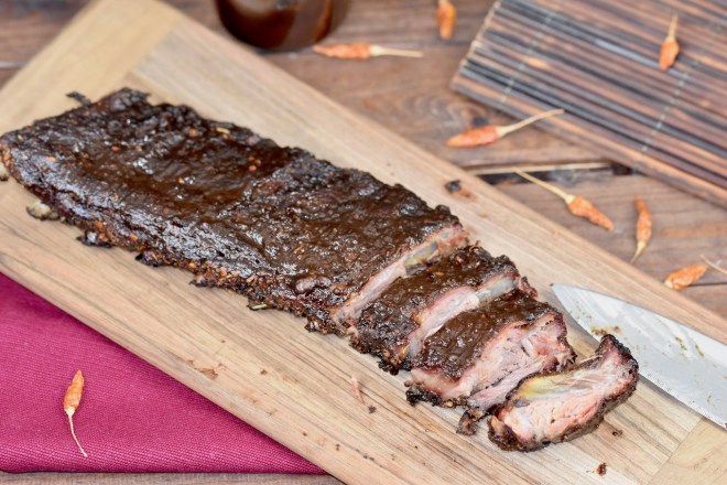 Ribs vom Holzkohlegrill auf Holzbrett. Ein Teil ist angeschnitten. Hintergrund dunkel.