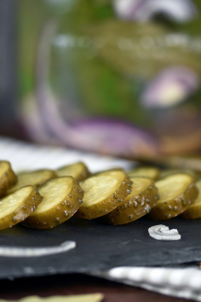 Fermentierte Gurken in einem Einmachglas. Davor liegen die Senfgurken angeschnitten auf einer Schieferplatte.