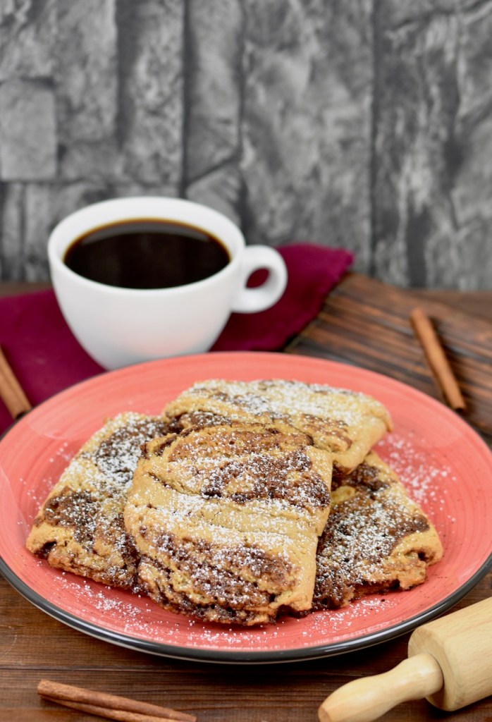 Glutenfreie Franzbrötchen auf rotem Teller. Im Hintergrund sieht man Zimtstangen, eine weiße Tasse gefüllt mit Kaffee und ein Stück von einem Nudelholz. Der Hintergrund ist dunkel.