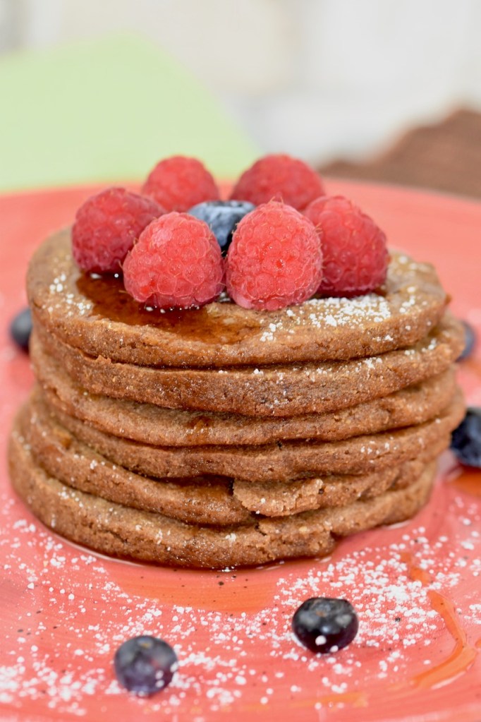 Glutenfreie Pfannkuchen gestapelt auf roten Teller, garniert mit Himbeeren, Blaubeeren, Ahornsirup und Puderzucker. Hintergrund hell. Nahaufnahme