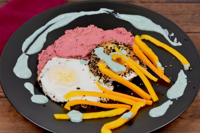 Knusprige Spiegeleier mit einer Kruste aus Sesam und Schwarzkümmelsamen mit pinkem Rote Beete Hummus und grüner Tahini-Sauce. Dazu gibt es gelbe Paprikastreifen Angerichtet ist alles auf einem schwarzen Teller.