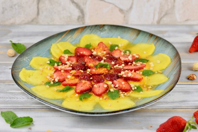 Erdbeer-Mango-Carpaccio mit Minze und Orangen-Dressing auf türkisem Teller garniert mit Minze und Haselnusskernen. Hintergrund weiß.