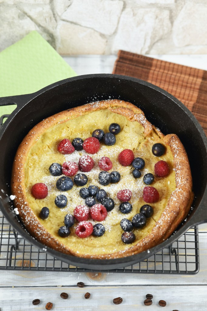 Glutenfreier Dutch Baby im schwarzen Gusseisentopf, dekoriert mit Himbeeren, Blaubeeren und Puderzucker. Hintergrund hell.