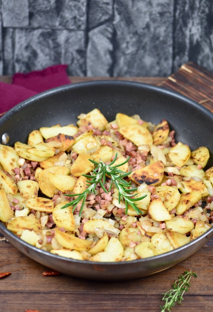 Knusprige Bratkartoffeln mit Speck und Zwiebeln in einer Pfanne, garniert mit Rosmarin