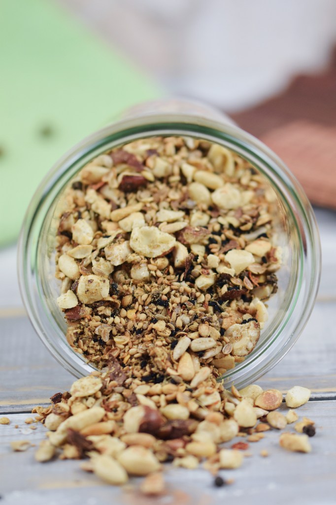 Dukkah selber gemacht in einem liegenden Glas. Etwas Dukkah ist herausgerutscht. Hintergrund hell.